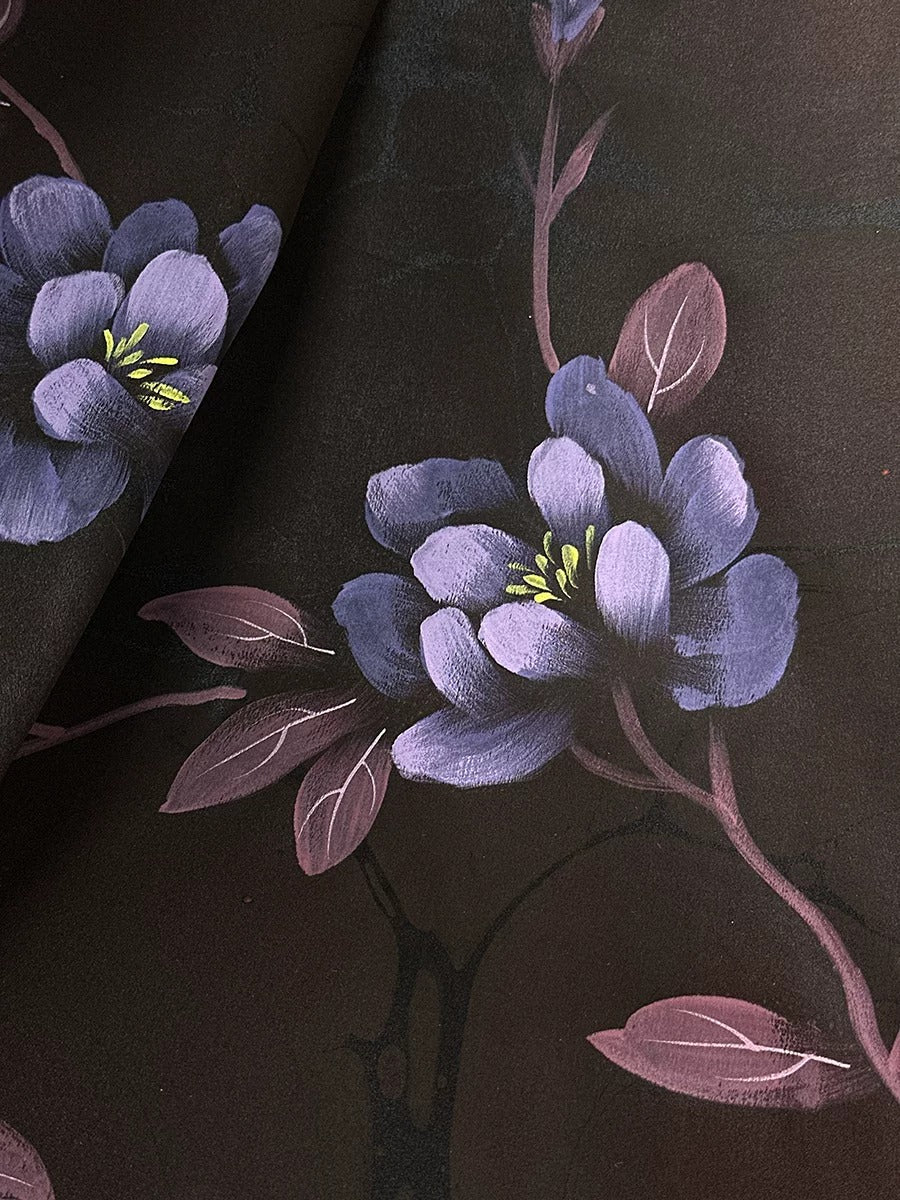 a close-up view of a dark brown fabric with a floral pattern. The pattern consists of purple flowers with green centers, along with leaves and stems. The fabric appears to be a piece of clothing or textile, possibly a garment or textile product.