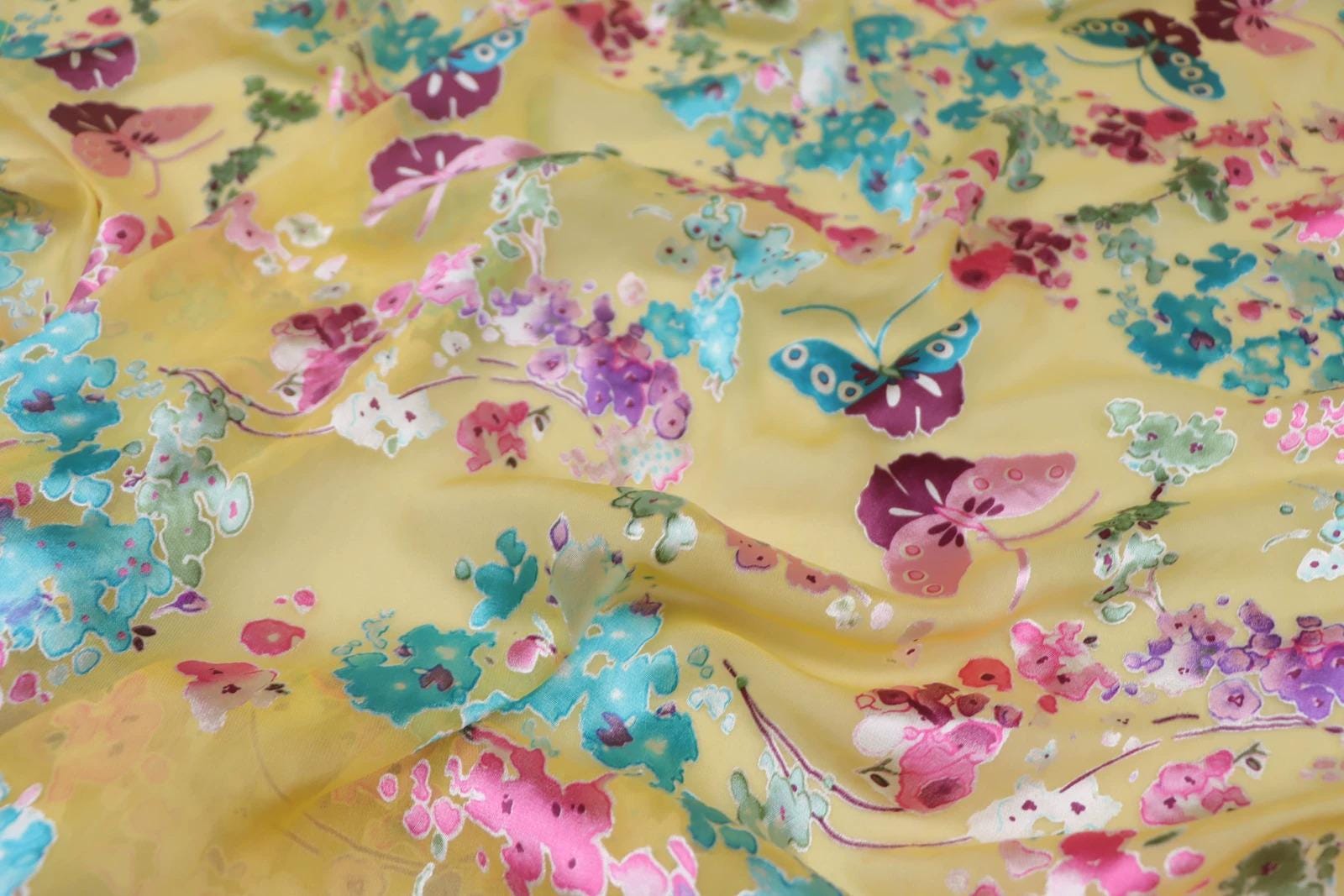 The image is a close-up of a fabric with a vibrant floral pattern in shades of yellow, pink, blue, and green. The pattern features various flowers and butterflies, creating a lively and colorful design.