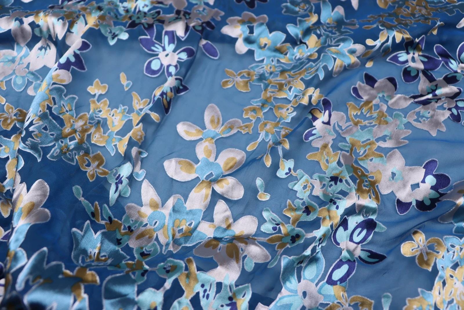 a close-up view of a fabric with a vibrant floral pattern in shades of blue, white, and gold. The pattern is intricate and detailed, with numerous small flowers and leaves scattered across the fabric. The fabric appears to be made of silk or a similar material, and the colors are vivid and eye-catching.