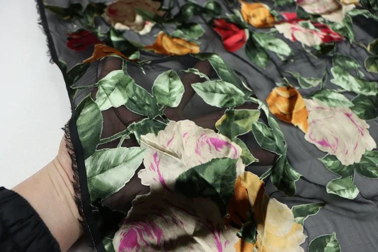 A close-up view of a hand holding a fabric with a floral pattern, featuring a mix of red, orange, and white flowers against a dark background.