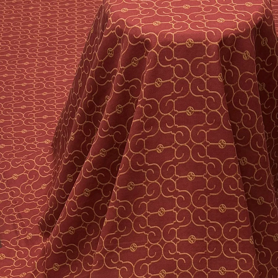 a close-up view of a red fabric with a repeating pattern of gold swirls and circles. The fabric appears to be folded or draped, creating a visually striking and intricate design.