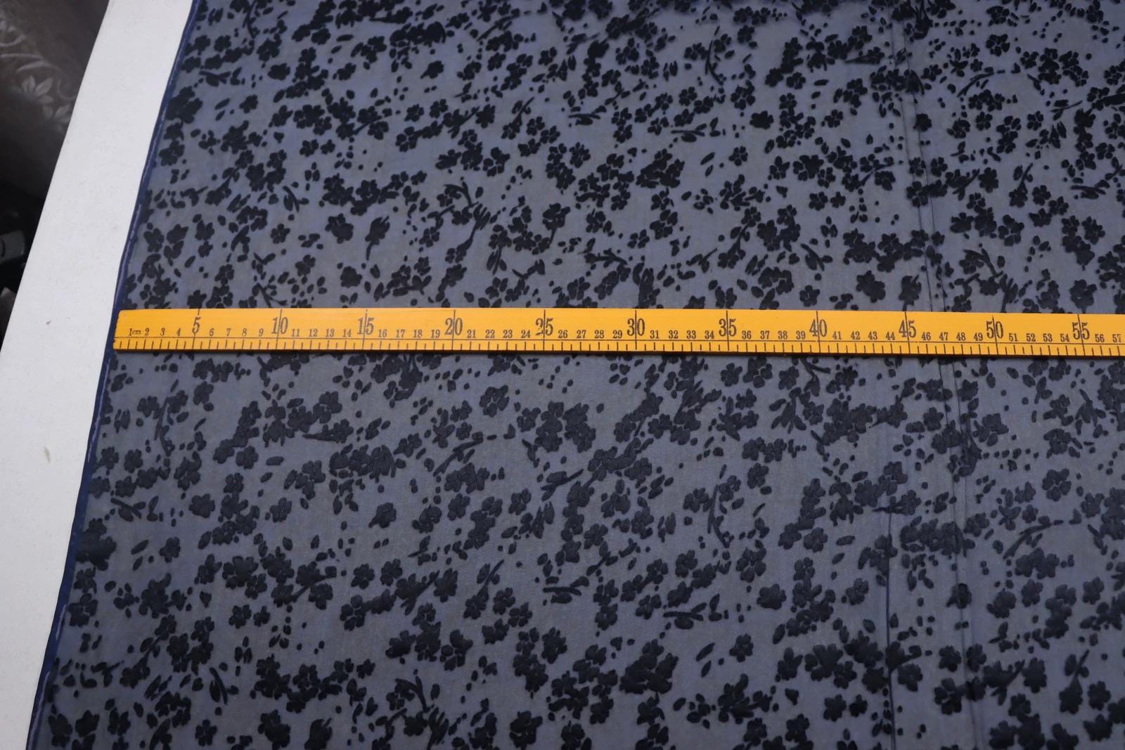 a close-up view of a fabric with a black floral pattern, and a yellow measuring tape is placed across the fabric, indicating the size or dimensions of the fabric.