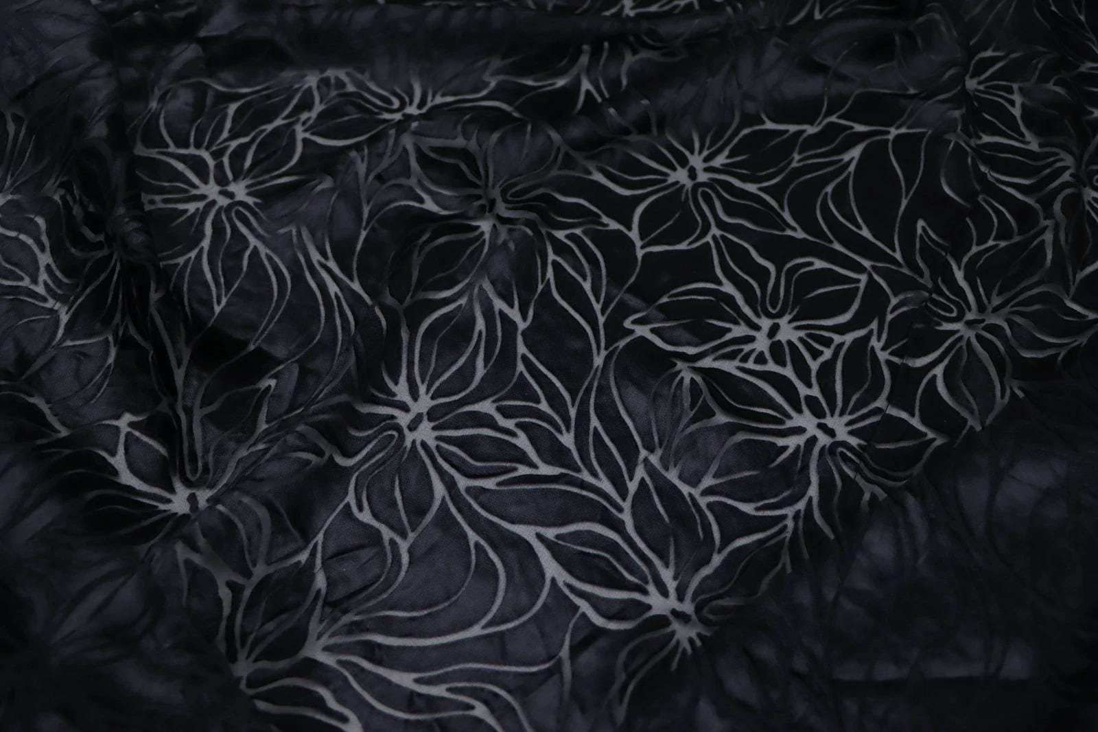 a close-up view of a dark fabric with a repeating floral pattern. The pattern consists of white flowers with black outlines, creating a visually striking contrast against the dark background.