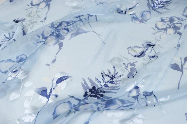 a close-up view of a fabric with a floral pattern in shades of blue and white. The fabric appears to be light blue, and the pattern consists of various blue flowers and leaves. The fabric is crumpled, giving it a soft and delicate appearance.