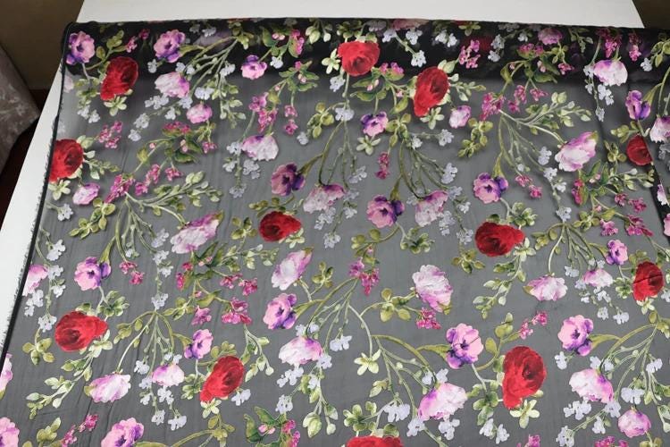 a close-up view of a fabric with a floral pattern. The fabric is predominantly black, but it is adorned with a variety of colorful flowers, including red, pink, and white flowers, as well as green leaves. The flowers are arranged in a repeating pattern across the fabric, creating a visually striking and intricate design.