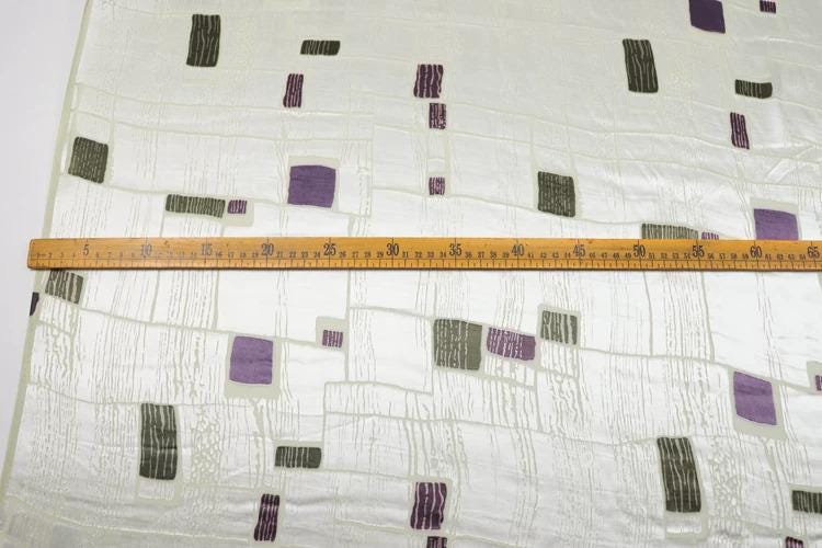 a close-up view of a fabric with a pattern of squares and rectangles in various shades of purple and green. A ruler is placed horizontally across the fabric, indicating the size or dimensions of the fabric.