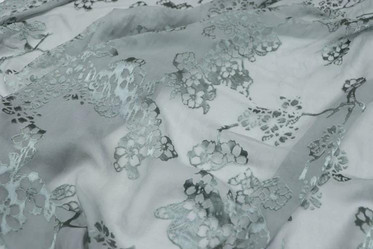 a close-up view of a fabric with a floral pattern, featuring a repeating design of small flowers or leaves in shades of gray and white.