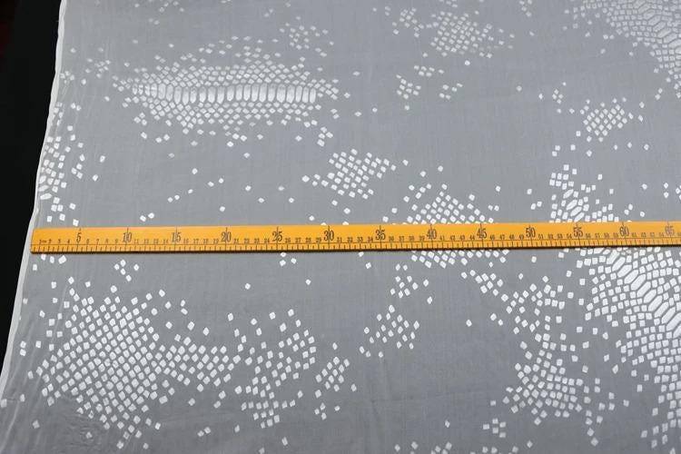 a gray fabric with a pattern of small white squares or dots on it, and a yellow measuring tape is placed across the fabric, indicating the size or dimensions of the fabric.