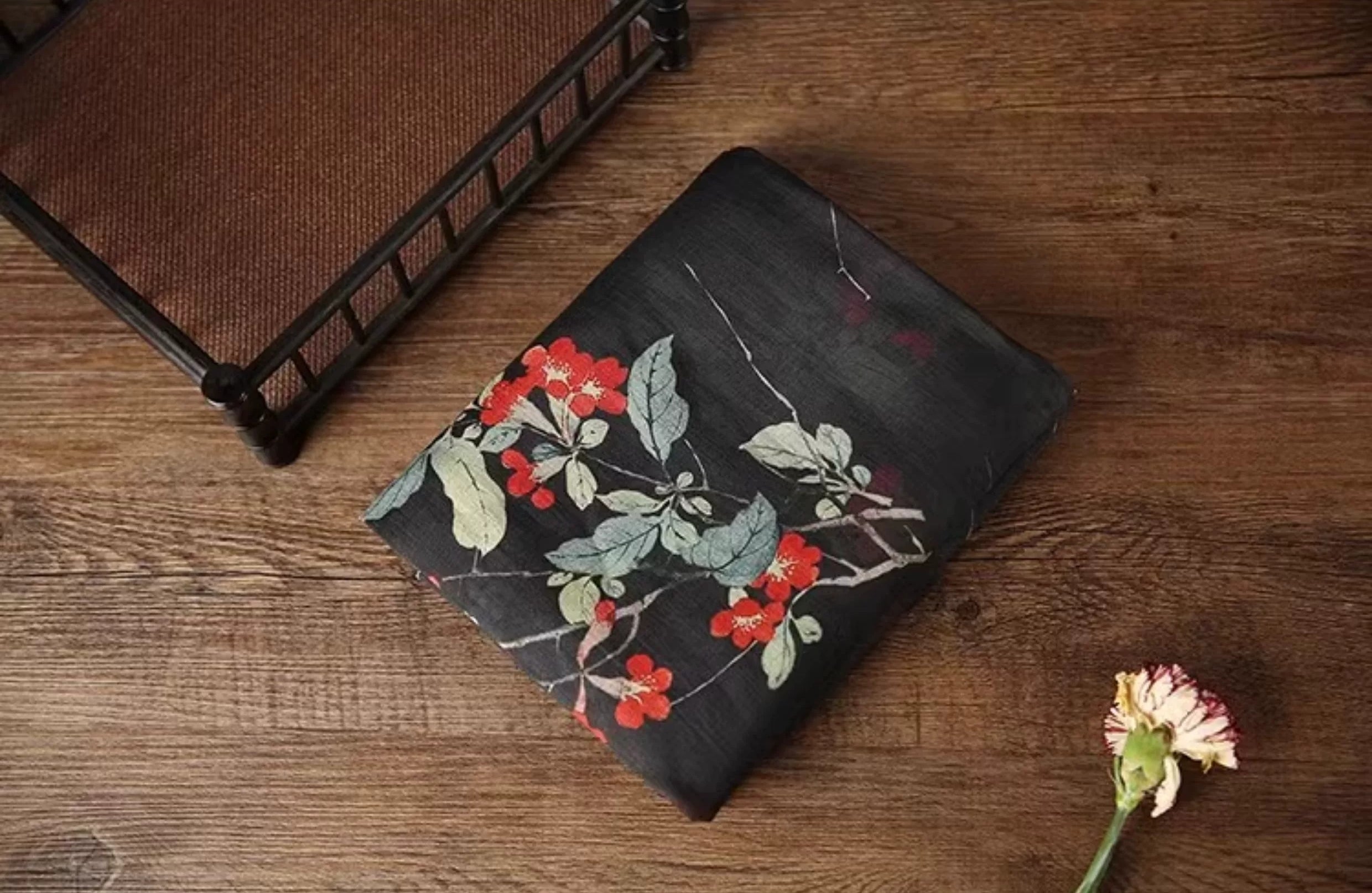 a black fabric with a floral pattern, including red and white flowers, resting on a wooden surface. There is also a small white flower nearby.