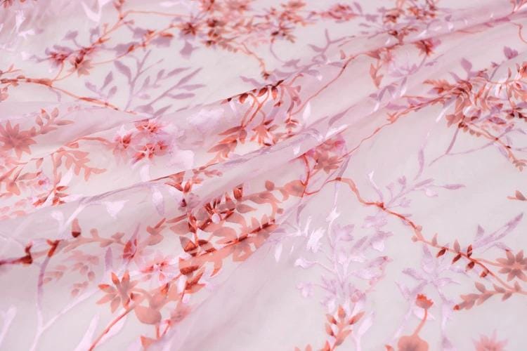 a close-up view of a fabric with a pink floral pattern. The fabric appears to be made of sheer material, with delicate pink flowers and leaves visible on the surface. The pattern is intricate and detailed, showcasing the craftsmanship of the fabric.