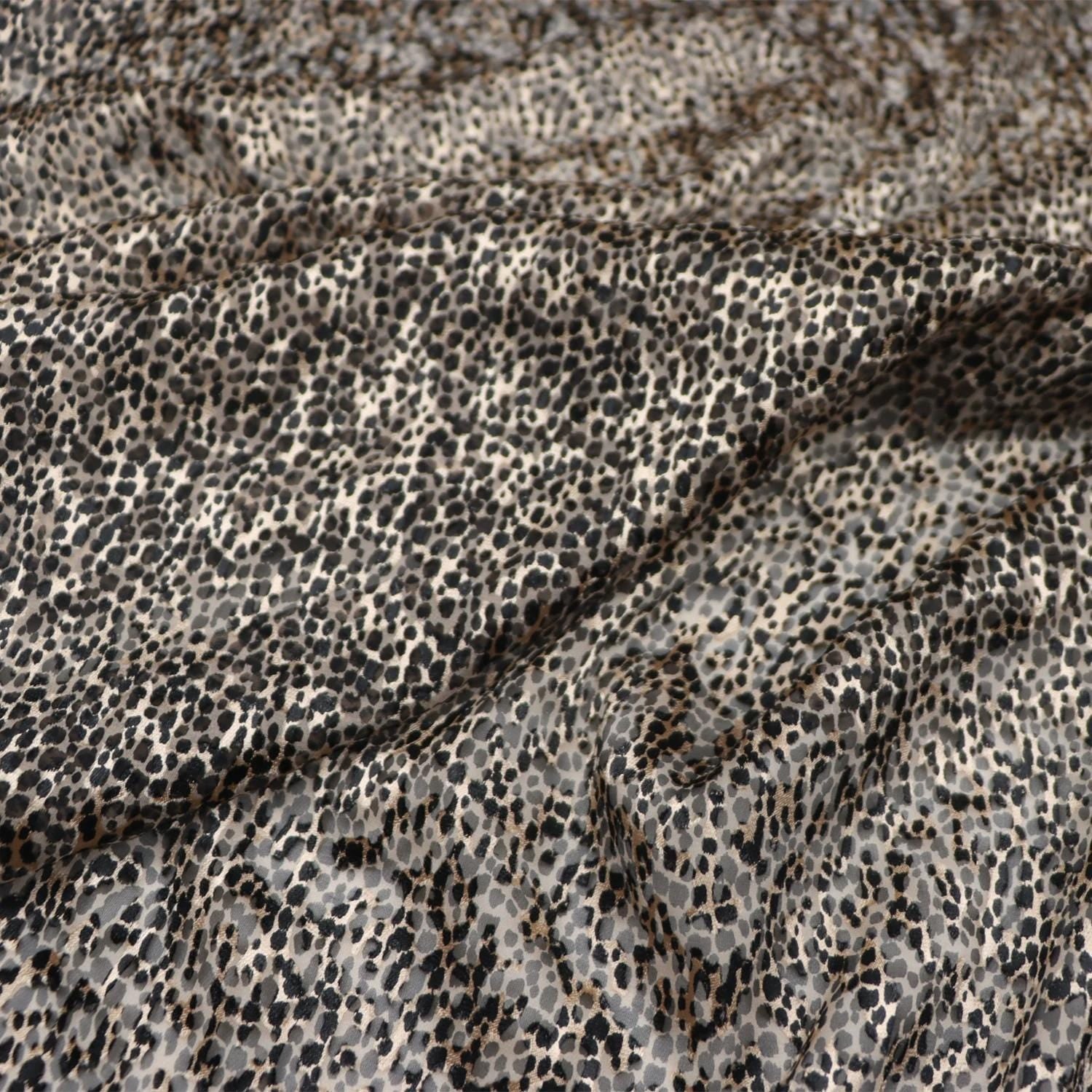a close-up view of a fabric with a distinctive leopard print pattern. The fabric appears to be made of a material with a textured surface, resembling the appearance of fur or scales. The pattern consists of small, irregularly shaped spots in various shades of brown and beige, creating a visually striking and intricate design.
