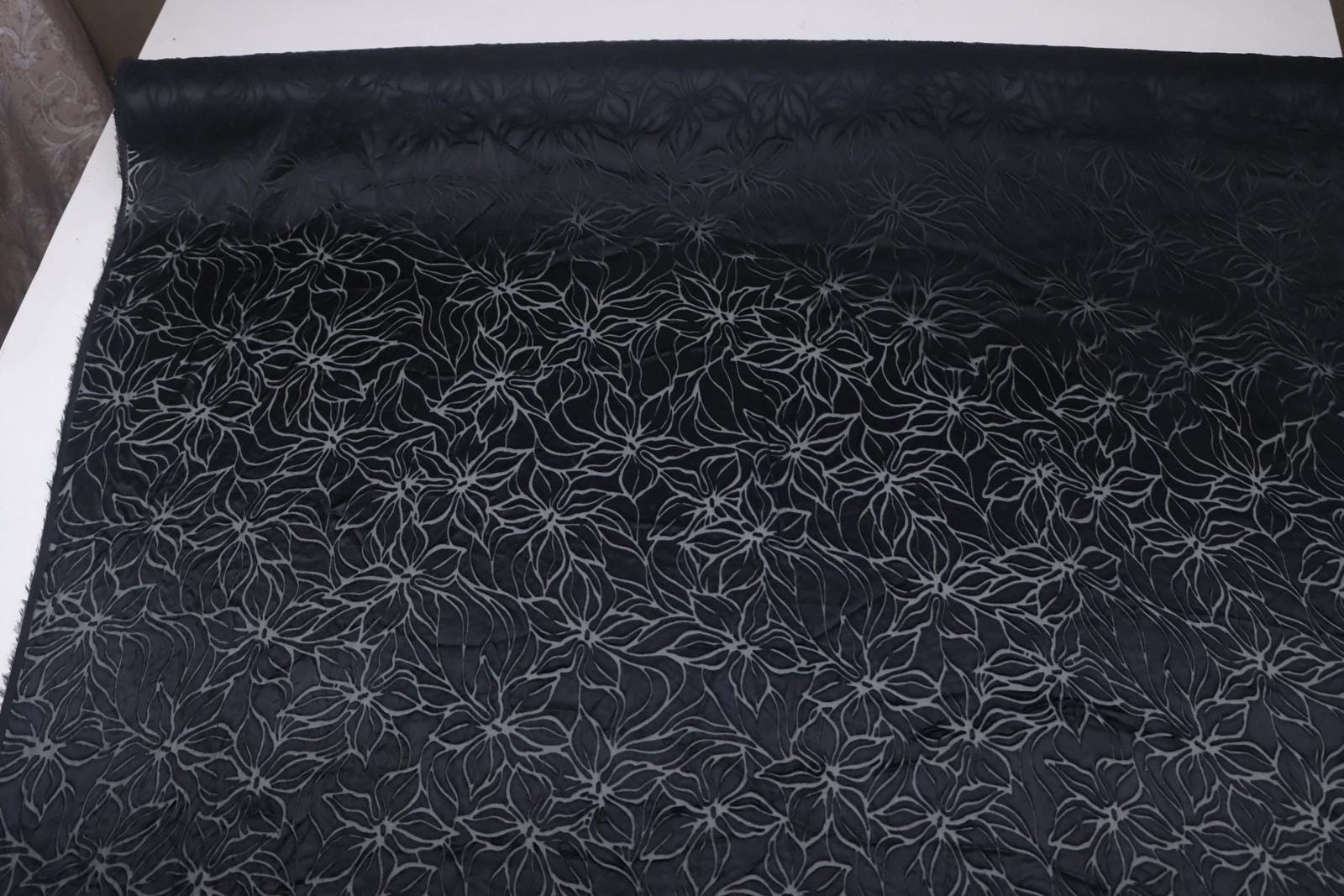 a close-up view of a black fabric with a repeating floral pattern. The pattern consists of white flowers arranged in a repeating design across the fabric's surface. The fabric appears to be a type of material, possibly a textile or a piece of clothing, with a dark background that contrasts with the white floral pattern.