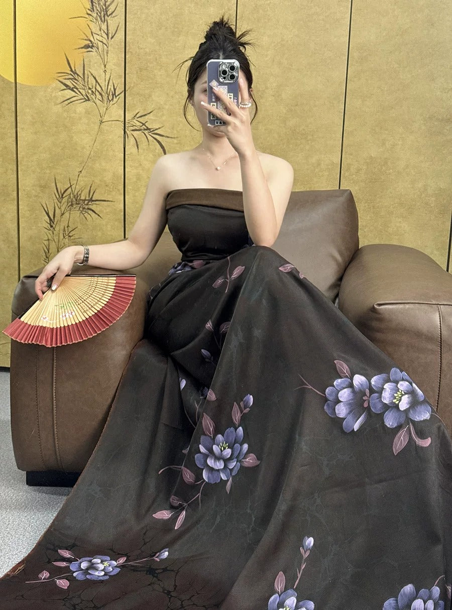 A woman in a long black dress with purple flowers is sitting on a brown leather couch, holding a fan and taking a selfie with her phone.