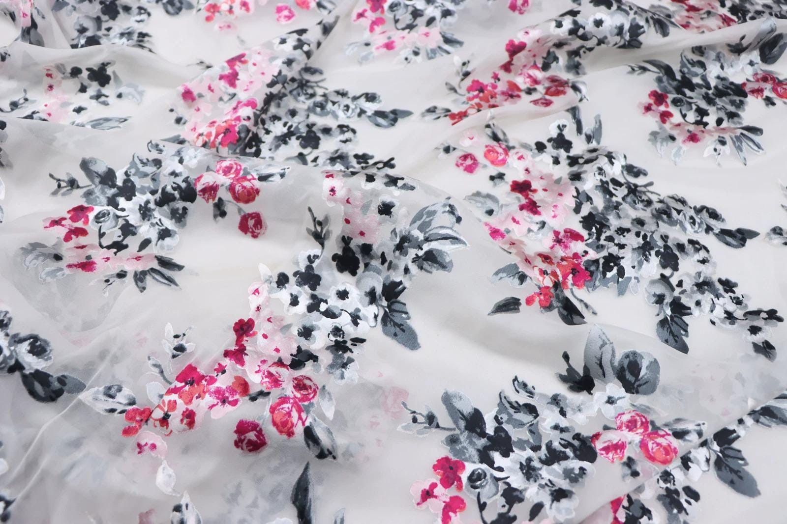 a close-up view of a fabric with a floral pattern in shades of pink and black. The pattern consists of various flowers and leaves, creating a visually appealing and intricate design. The fabric appears to be made of a soft, sheer material, which adds to the delicate and elegant appearance of the pattern.