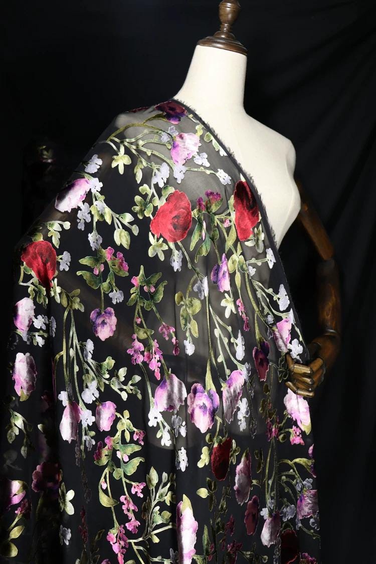 a mannequin wearing a black dress with a floral print. The dress has a sheer black fabric overlay, and the floral pattern includes red, pink, and white flowers. The mannequin is positioned against a black background, which highlights the details of the dress and the floral print.