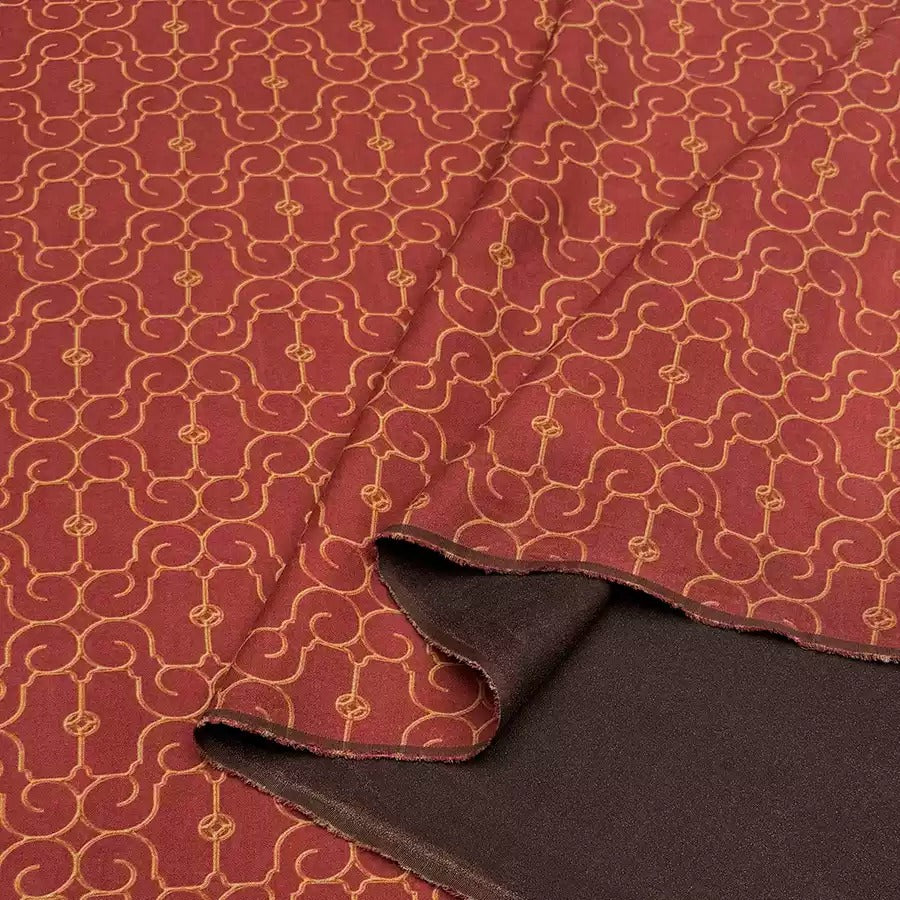 a close-up view of a fabric with a repeating pattern of red and gold designs. The fabric appears to be made of a material with a rich, deep red color and a golden, intricate pattern. The fabric is folded and partially unrolled, revealing the intricate design underneath.