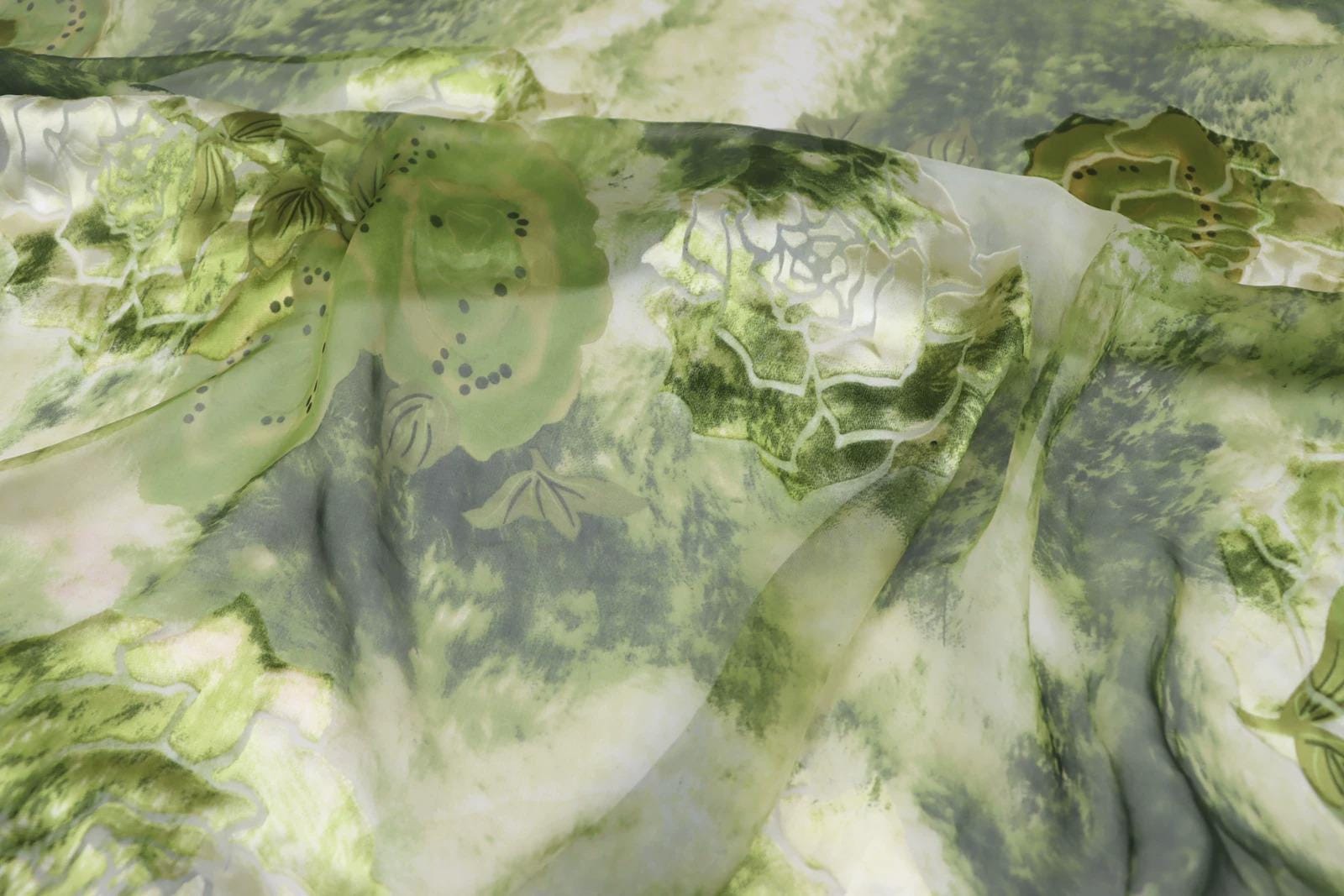 The image is a close-up of a fabric with a green and white floral pattern. The fabric appears to be made of silk or a similar material, and it has a slightly wrinkled texture. The pattern consists of various shades of green and white, creating a visually appealing and intricate design.
