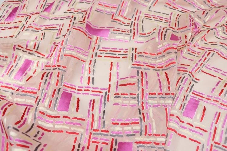 a close-up view of a fabric with a geometric pattern in shades of pink, red, and gray. The pattern consists of various shapes and lines, creating a visually striking and intricate design.