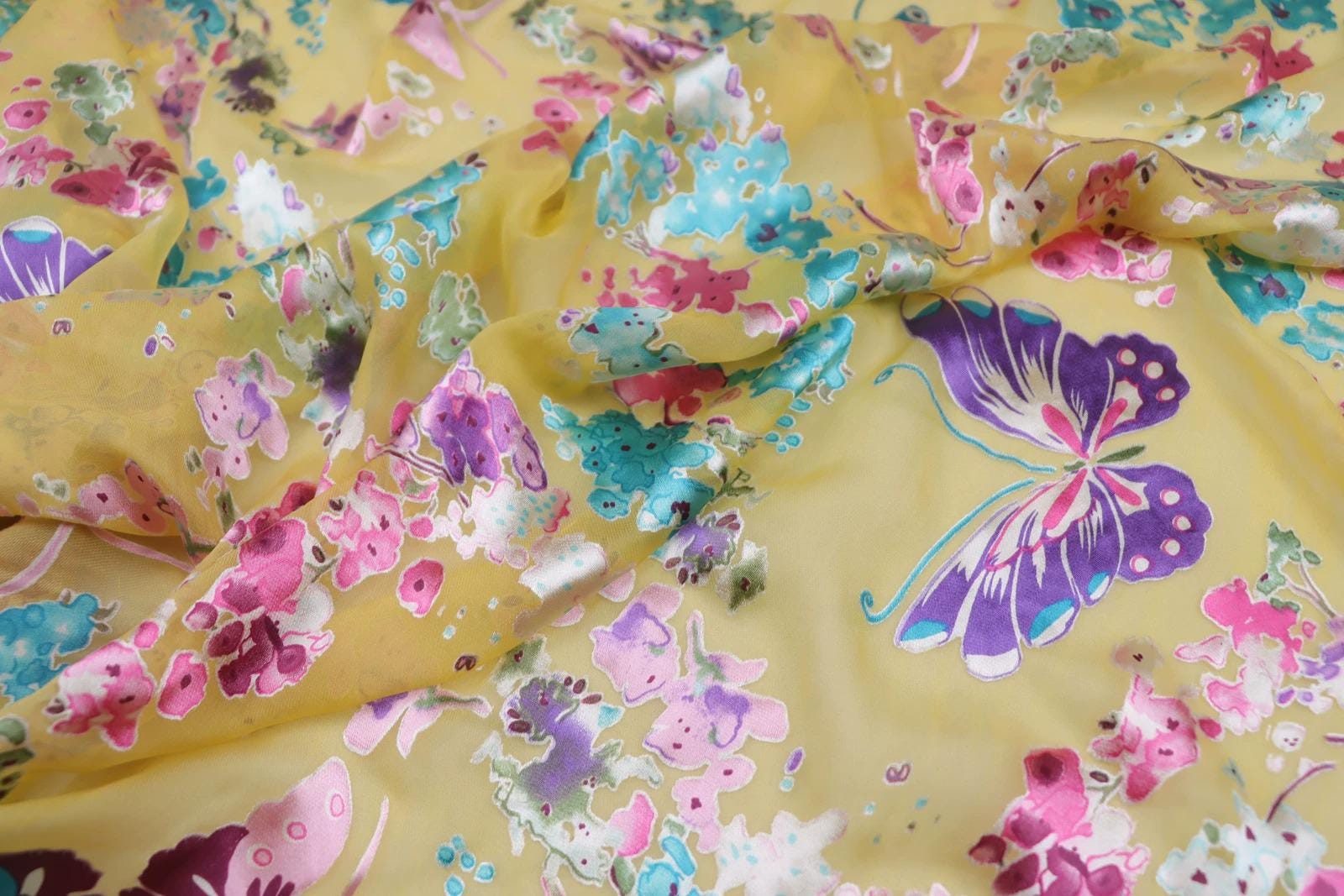 The image is a close-up view of a fabric with a vibrant, colorful butterfly pattern. The butterfly is the main focal point, with various shades of yellow, pink, and blue creating a lively and eye-catching design.