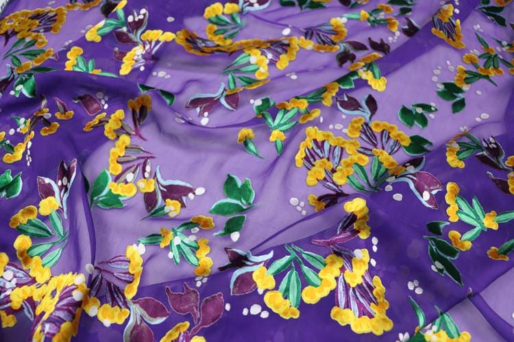 a close-up view of a fabric with a vibrant floral pattern in shades of purple, yellow, and green. The pattern features various flowers and leaves, creating a visually striking and colorful design.