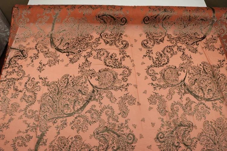a close-up view of a fabric with a repeating paisley pattern in shades of orange and black. The pattern is intricate and detailed, with various shapes and designs that create a visually striking and textured appearance.