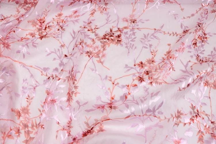 a close-up view of a fabric with a pink floral pattern. The fabric appears to be made of sheer material, with delicate pink flowers and leaves covering the entire surface. The pattern is intricate and detailed, showcasing the craftsmanship of the fabric.