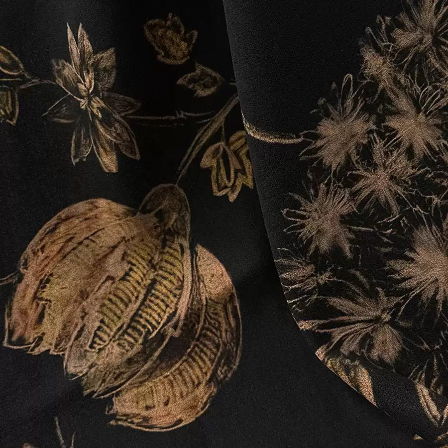 a close-up view of a black fabric with a floral pattern, including a large flower and smaller flowers. The fabric appears to be made of a shiny material, possibly silk or satin, and has a dark background.