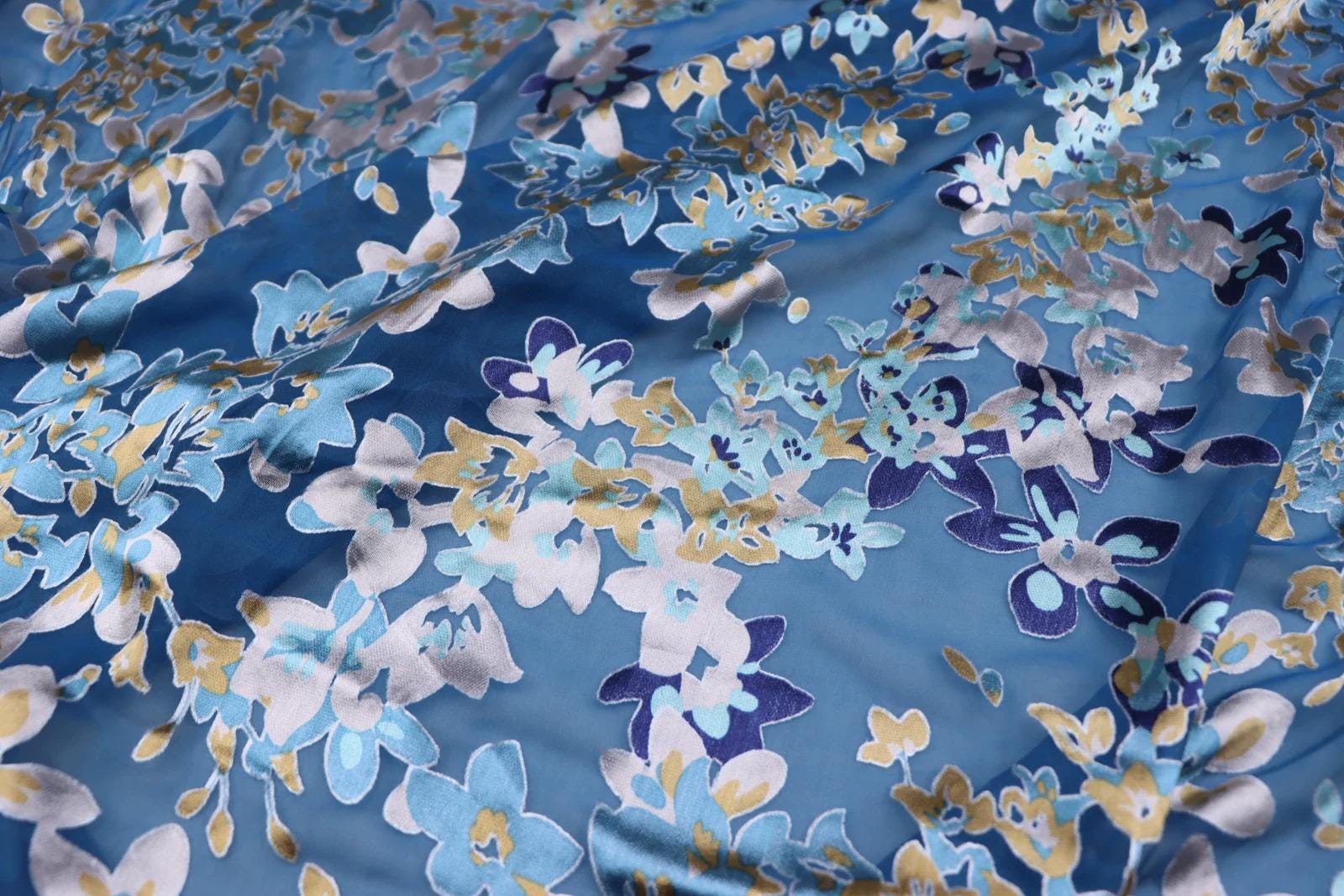 a close-up view of a fabric with a floral pattern in shades of blue, white, and gold. The fabric appears to be made of silk or a similar material, and the pattern is intricate and detailed.
