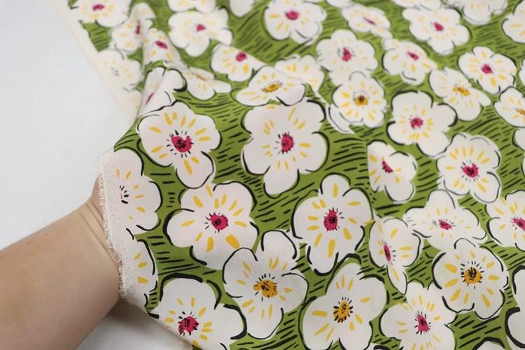 a close-up view of a hand holding a fabric with a repeating pattern of white flowers with yellow centers against a green background.