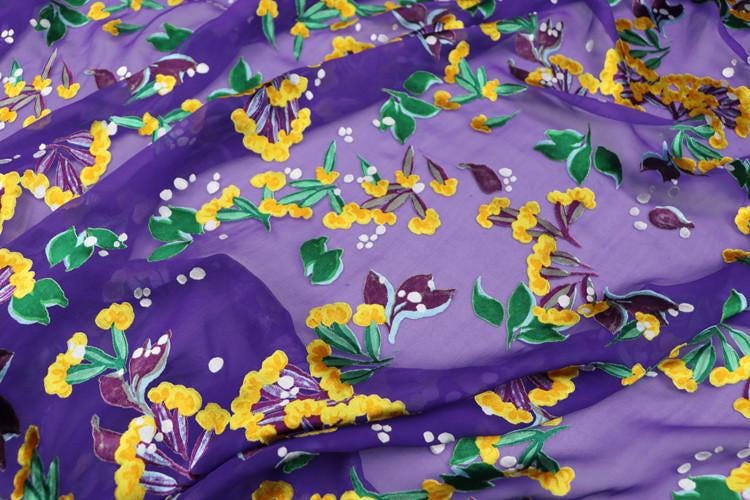 a close-up view of a fabric with a vibrant floral pattern in shades of purple and yellow. The pattern features various flowers and leaves, creating a lively and colorful design.