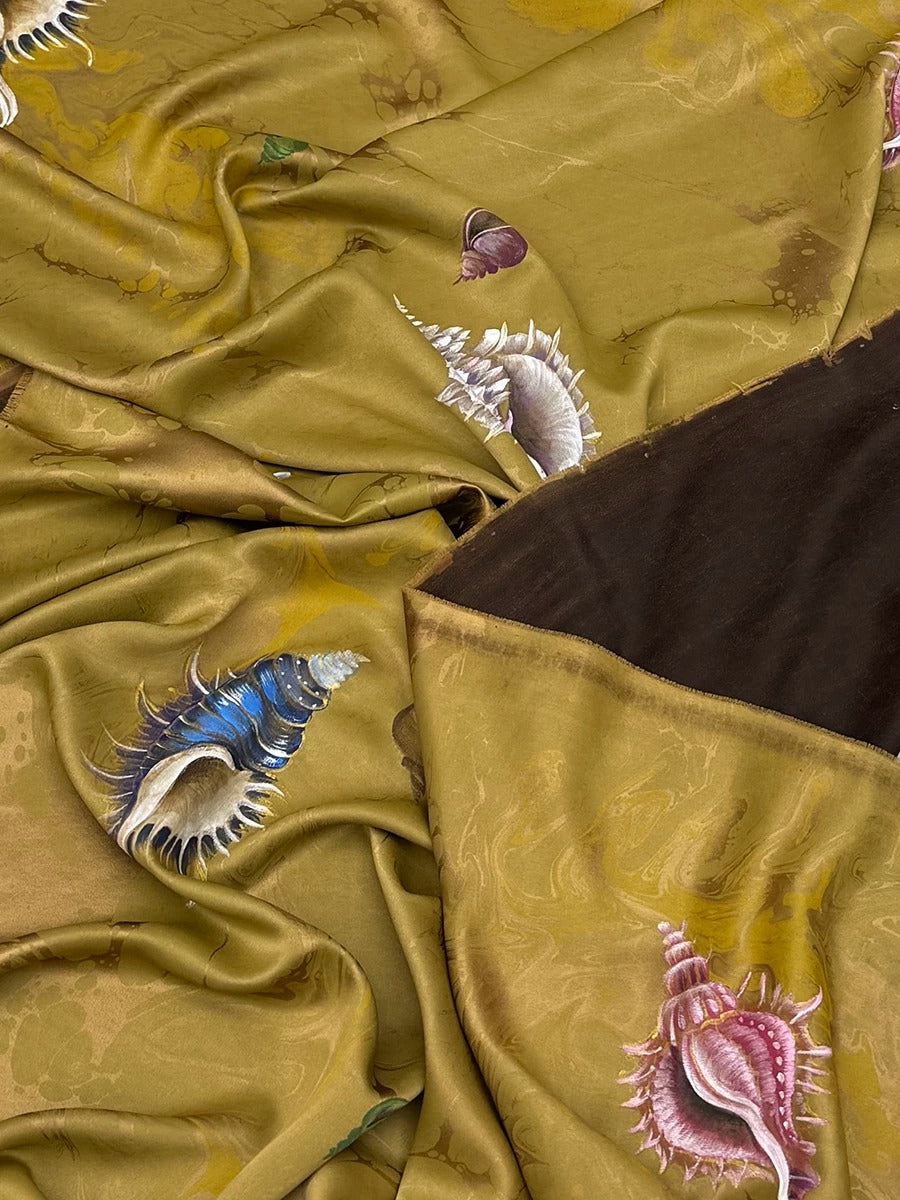 a close-up view of a fabric with a vibrant, colorful pattern of seashells and other marine life. The fabric appears to be made of silk or a similar material, with a rich gold-colored background. The seashells are depicted in various shades of blue, pink, and white, creating a visually striking and intricate design. The fabric is crumpled, adding a sense of texture and depth to the image.