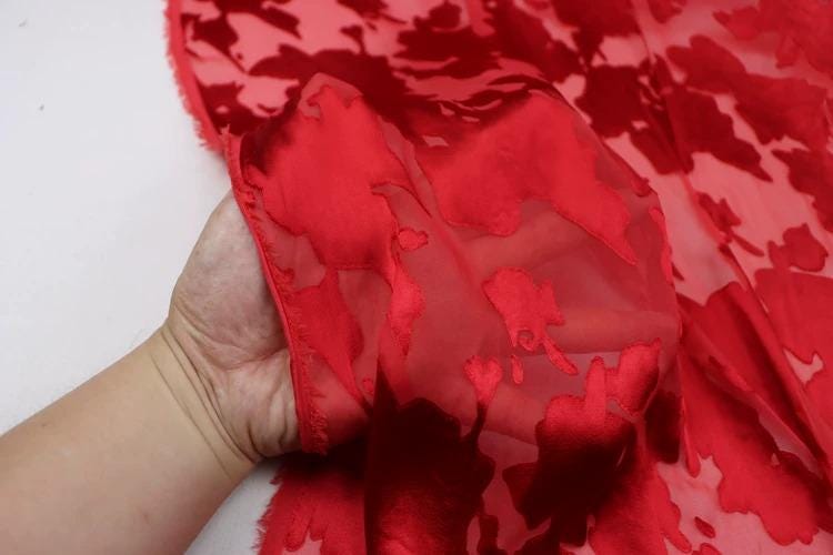 A hand is holding a piece of red fabric with a floral pattern, which appears to be a close-up view of the fabric.