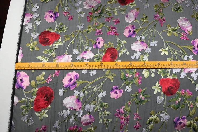 a close-up view of a fabric with a floral pattern, featuring red and pink flowers along with green leaves. A yellow measuring tape is placed across the fabric, likely for size reference or to measure the length of the fabric.