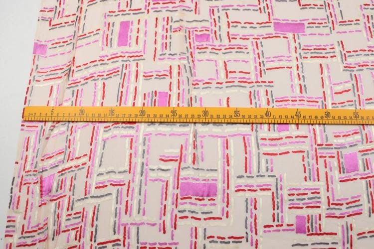 a close-up view of a fabric with a geometric pattern in shades of pink, red, and gray. A yellow measuring tape is laid across the fabric, providing a reference for size.