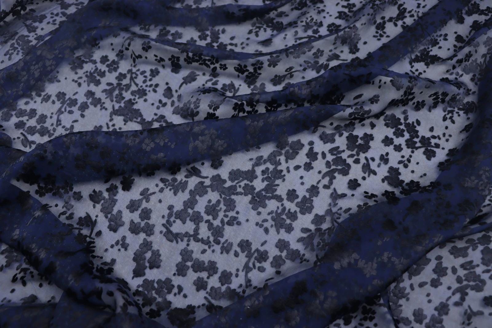 a close-up view of a fabric with a floral pattern, featuring a mix of blue and white colors. The fabric appears to be made of sheer material, with a dark blue color dominating the background.