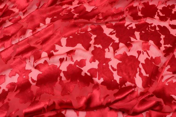 a close-up view of a fabric with a red floral pattern. The fabric appears to be made of a shiny, satin-like material, and the pattern consists of red flowers or leaves against a pink background. The fabric is crumpled and wrinkled, giving it a textured appearance.