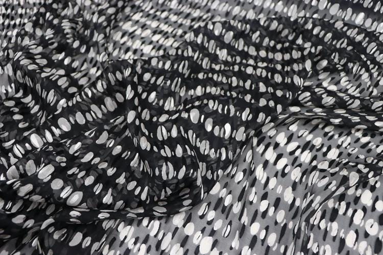 a close-up view of a fabric with a black and white polka dot pattern. The fabric appears to be made of a sheer material, with a crumpled texture. The pattern consists of numerous small white dots arranged in a diagonal pattern across the fabric. The image does not contain any text or other discernible objects or actions.