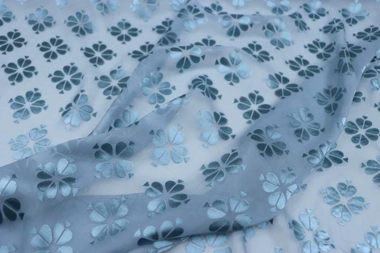 a close-up view of a fabric with a repeating pattern of blue and white flowers or leaves. The fabric appears to be made of a soft, flowing material, and the pattern is intricate and detailed.
