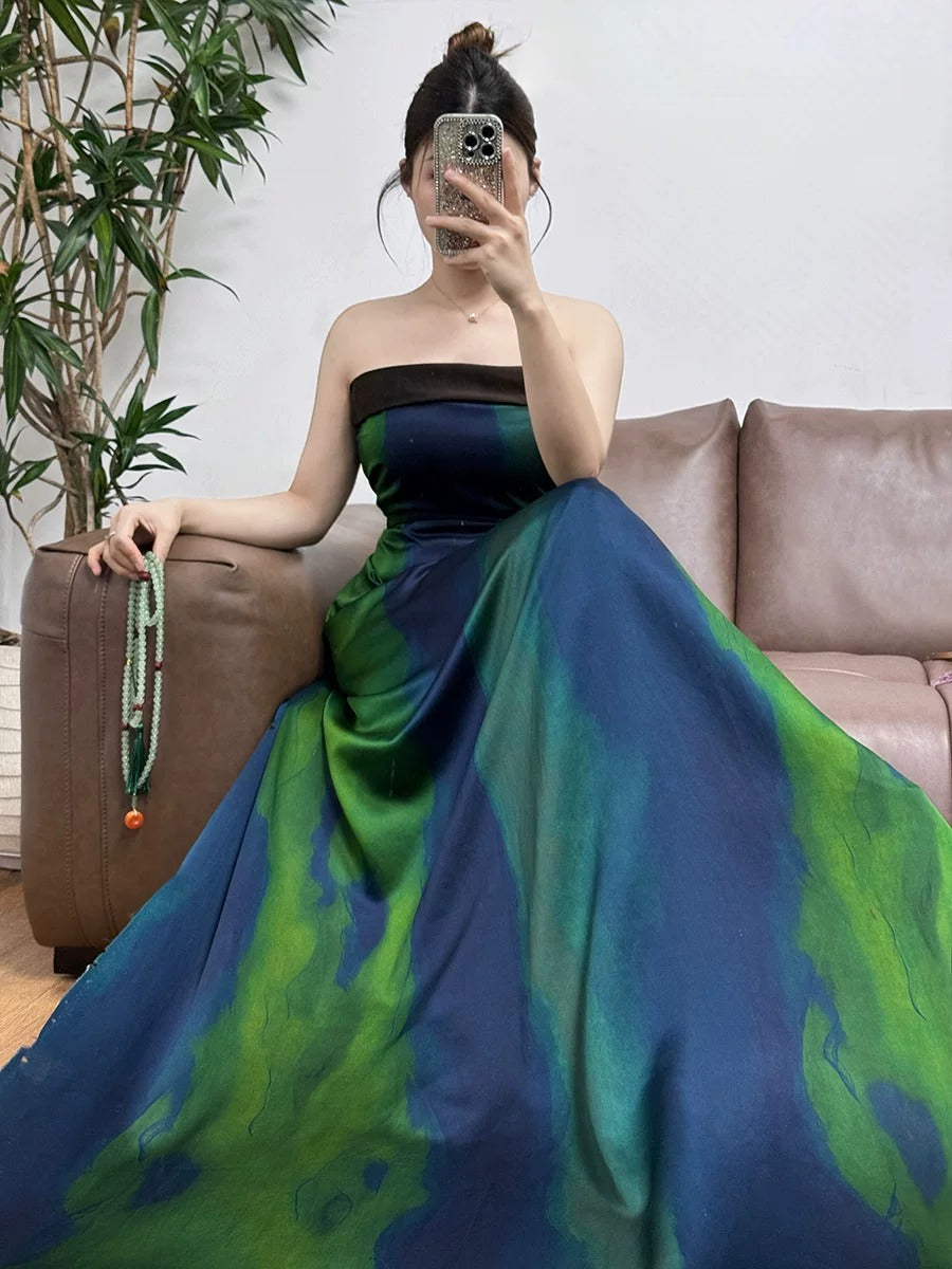 A woman in a long, colorful dress is sitting on a couch, taking a selfie with her phone.