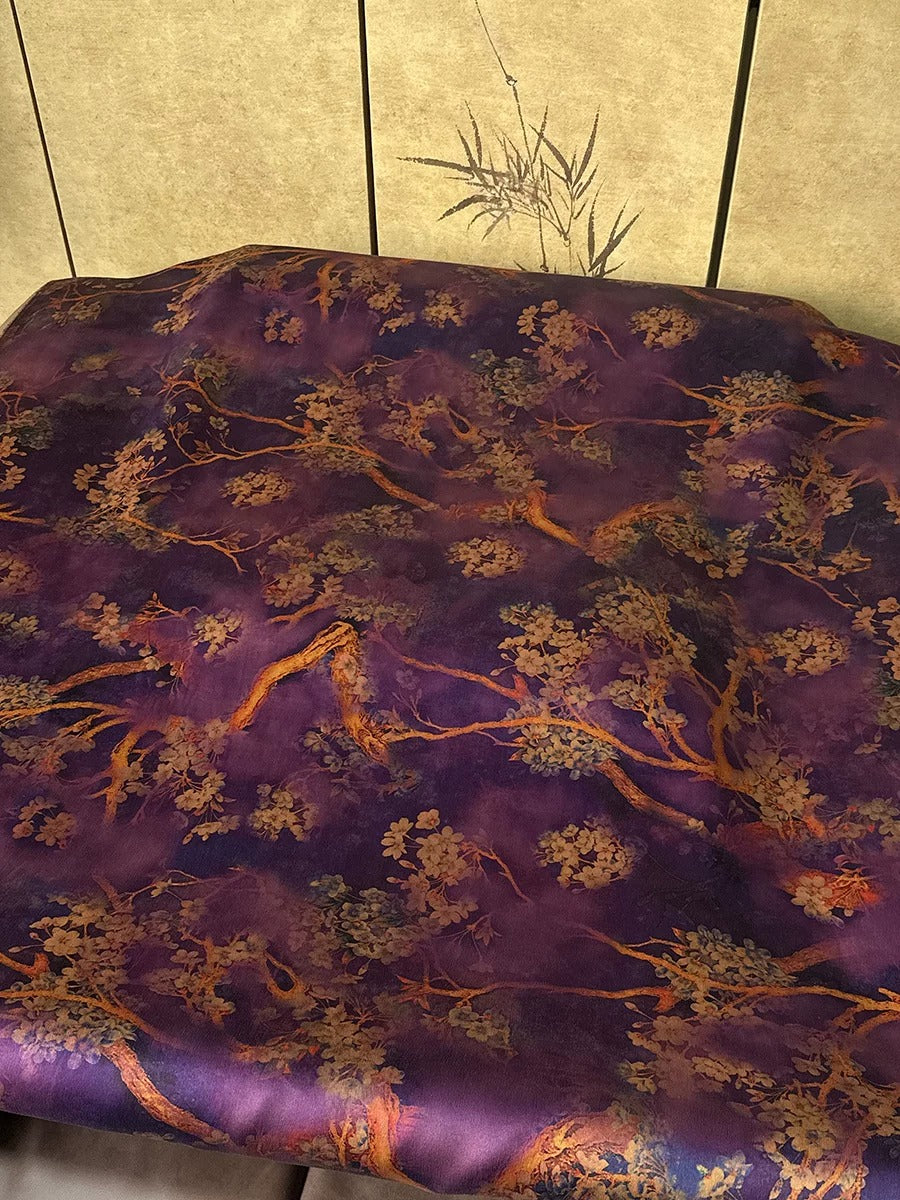 a close-up view of a fabric with a purple and gold floral pattern. The fabric appears to be a piece of clothing or textile, possibly a bedspread or pillowcase. The pattern is intricate and detailed, with the flowers and branches depicted in varying shades of purple and gold. The fabric is spread out on a surface, possibly a table or countertop, and there is a plant visible in the background.